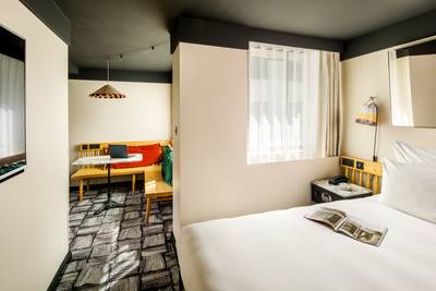 Modern hotel room with a cosy bed, a small table with a laptop, a sitting area, and a window with sheer curtains. Bright and inviting atmosphere.