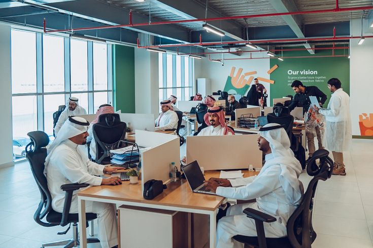 A modern office with people working at desks, some wearing traditional attire. Large windows, green wall with a vision statement. Collaborative environment.
