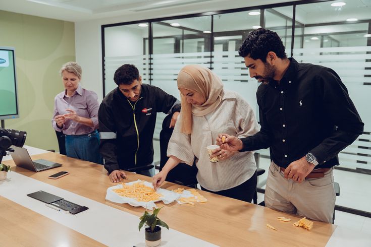 Four people in an office gathering around a table, sharing and enjoying French fries from a paper towel spread.