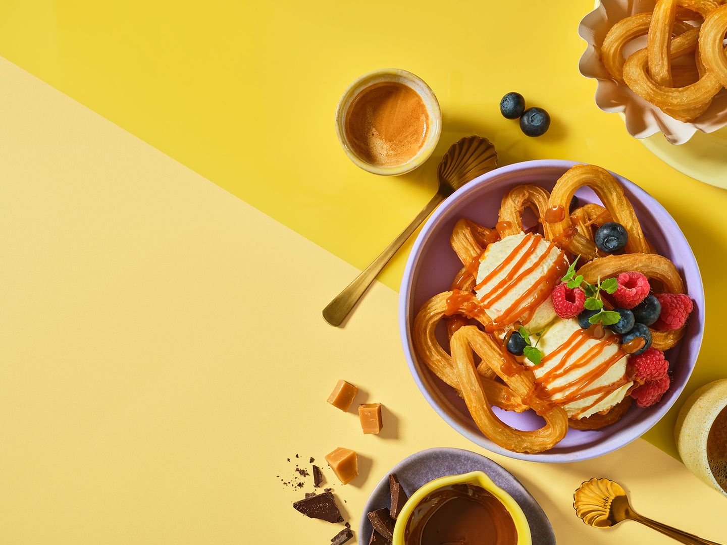 Churros with ice cream, caramel drizzle, raspberries, and blueberries in a purple bowl on a yellow background, with espresso and chocolate pieces nearby.