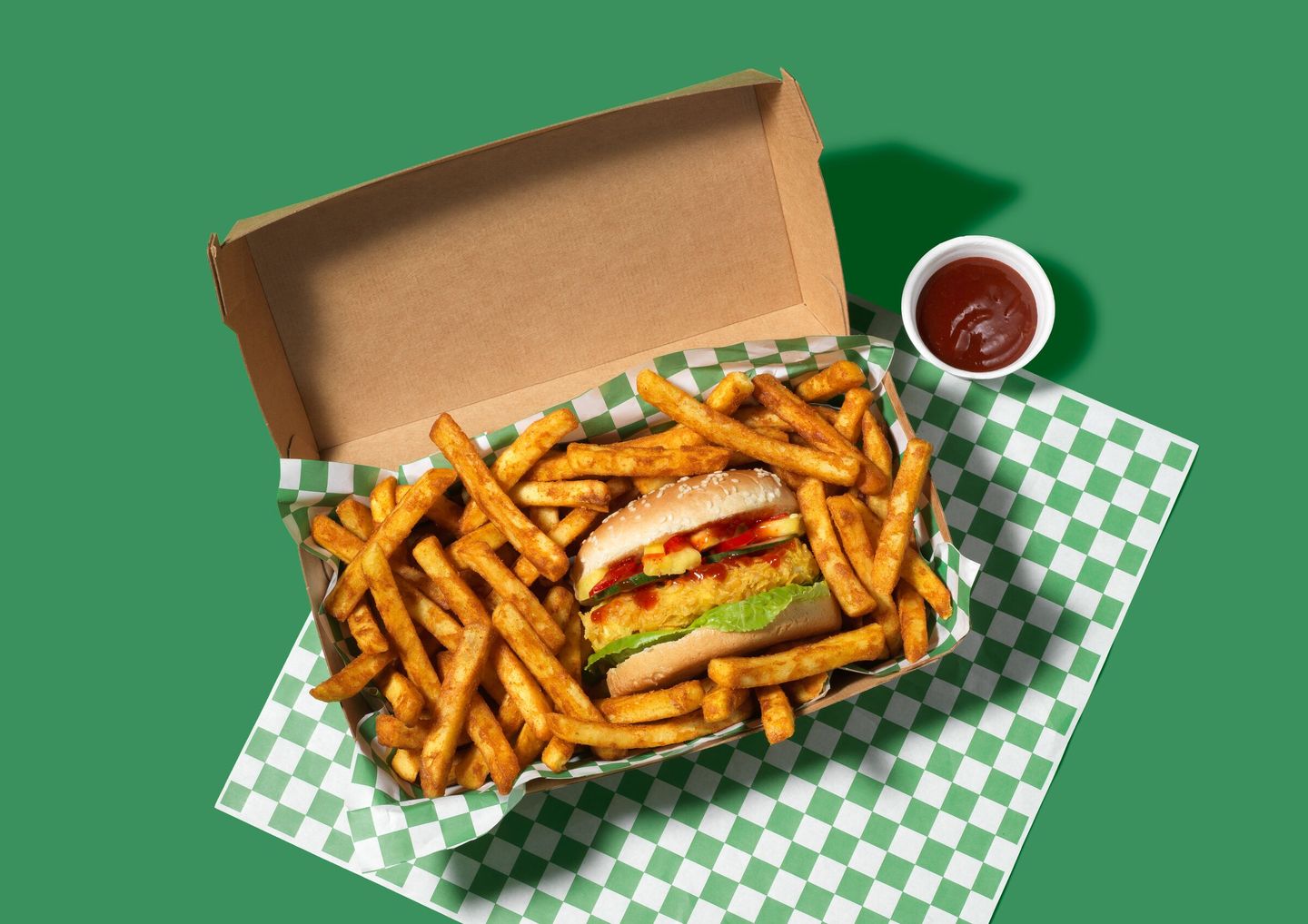 Chicken burger with lettuce and toppings in a box, surrounded by fries, on green checkered paper. Cup of ketchup on the side.