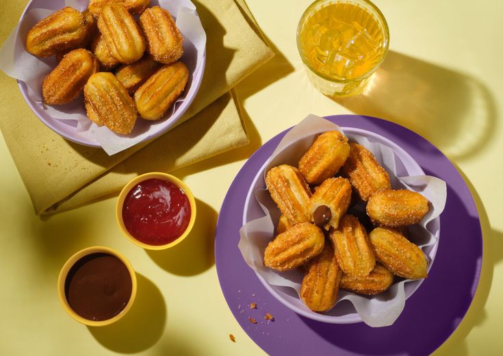 Bowls of mini churros on purple plates with chocolate and strawberry dips, set on a yellow surface with a glass of iced beverage.
