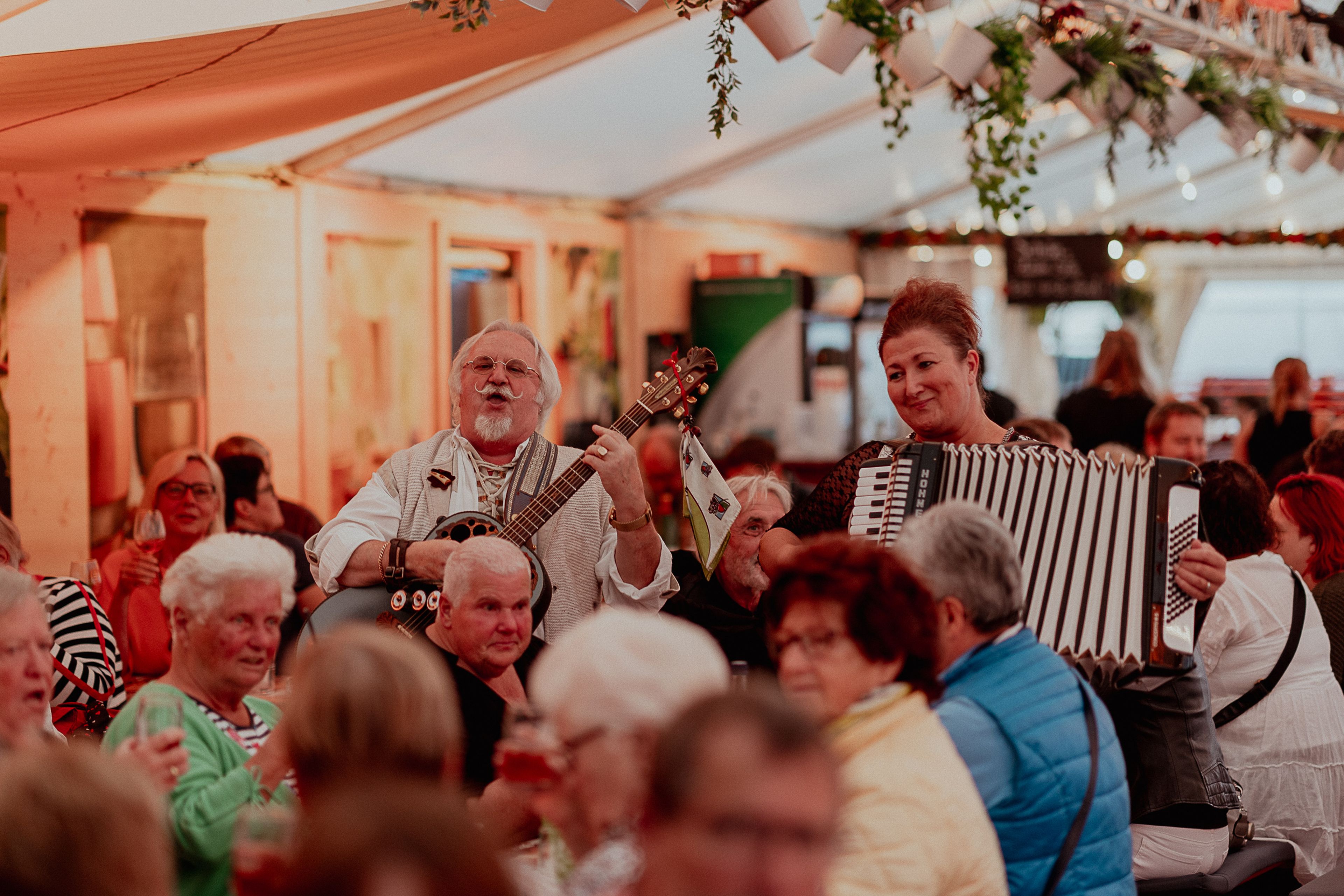 Musiker, die Gitarre und Akkordeon spielen, unterhalten eine lebhafte Menge in einem warm beleuchteten Zelt, das mit Menschen gefüllt ist, die an Tischen sitzen.