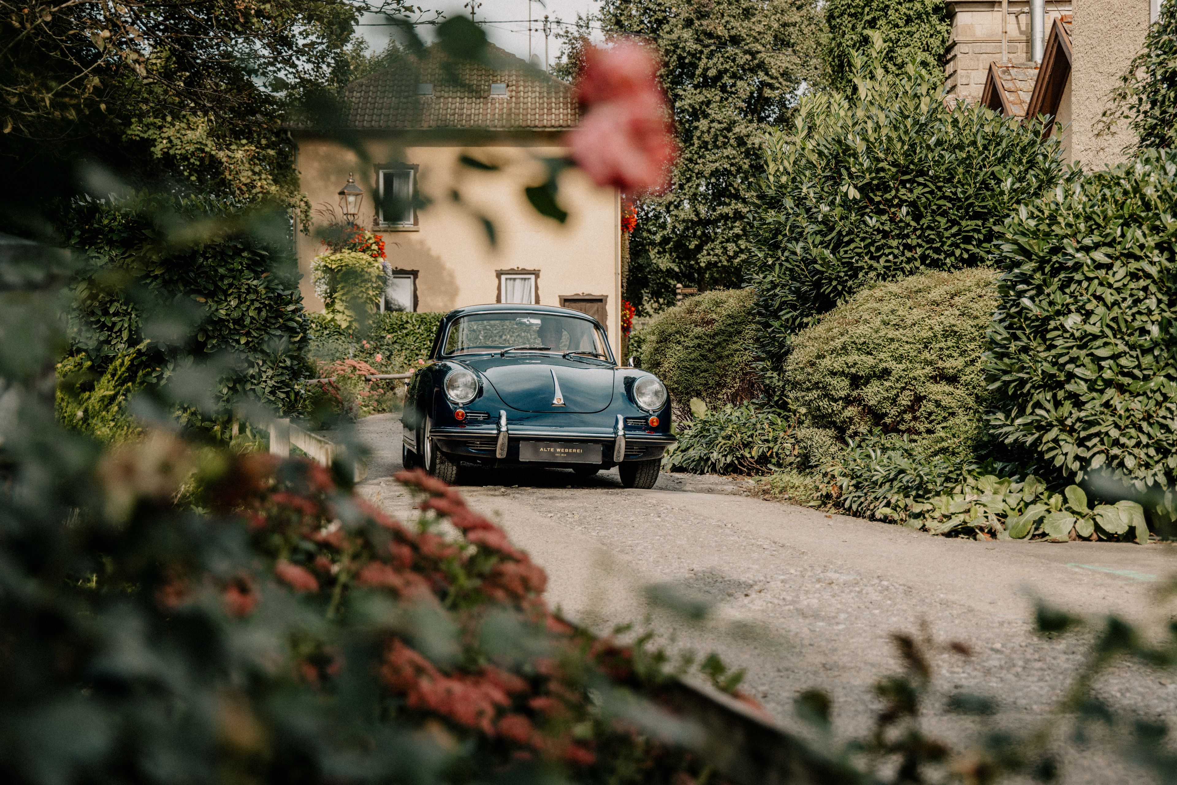 Oldtimer parkt auf einer schmalen, von Bäumen gesäumten Straße mit einem malerischen Haus im Hintergrund, umgeben von üppigem Grün und Blumen.