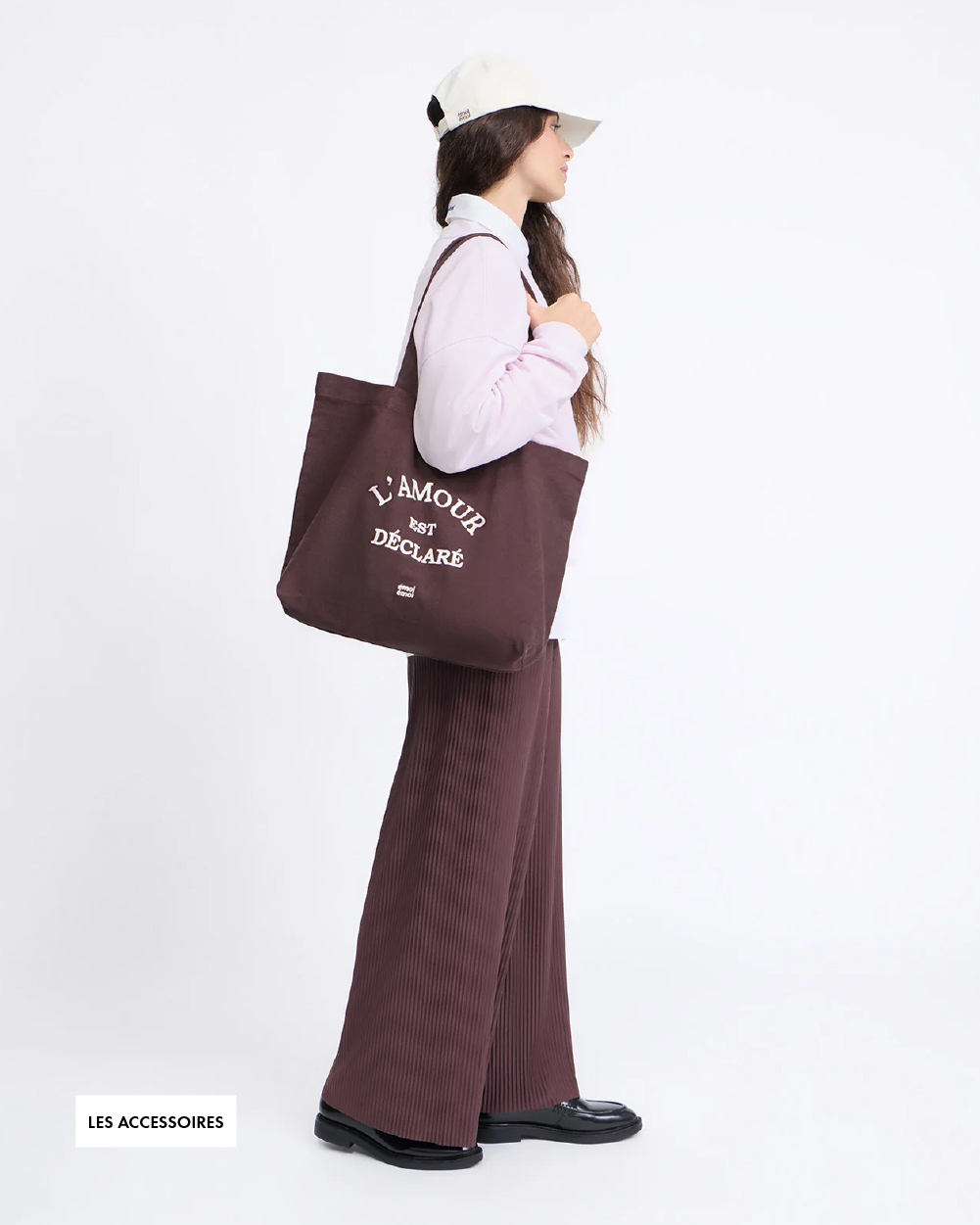 Woman in a white cap, pink sweater, and brown pleated pants holds a brown tote bag with "L