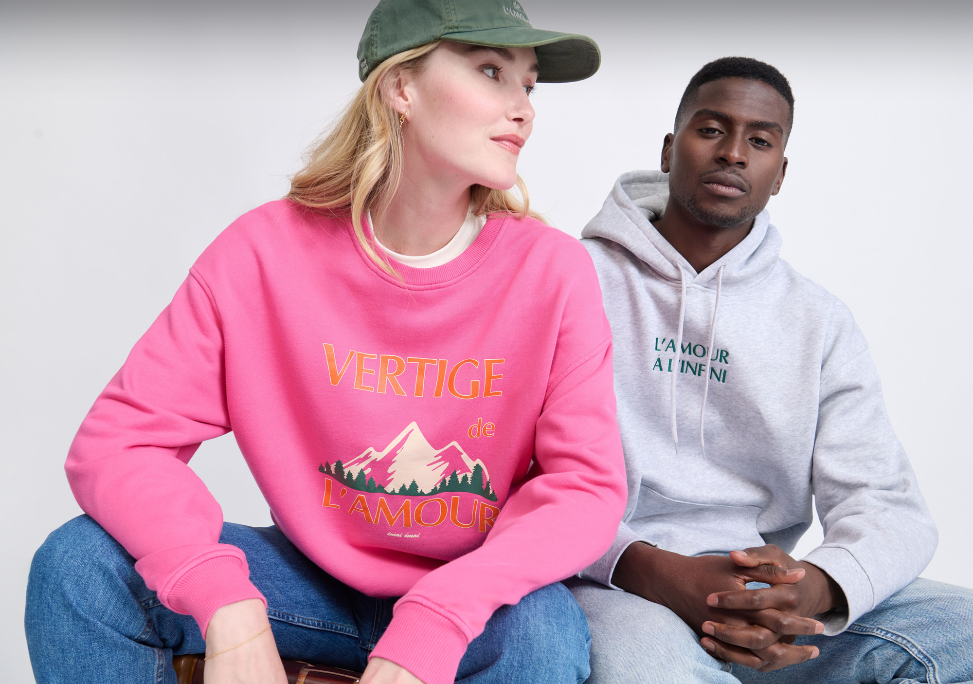 Two people sitting; one wears a pink sweatshirt with mountain design, the other a gray hoodie. Casual and relaxed pose against a simple backdrop.