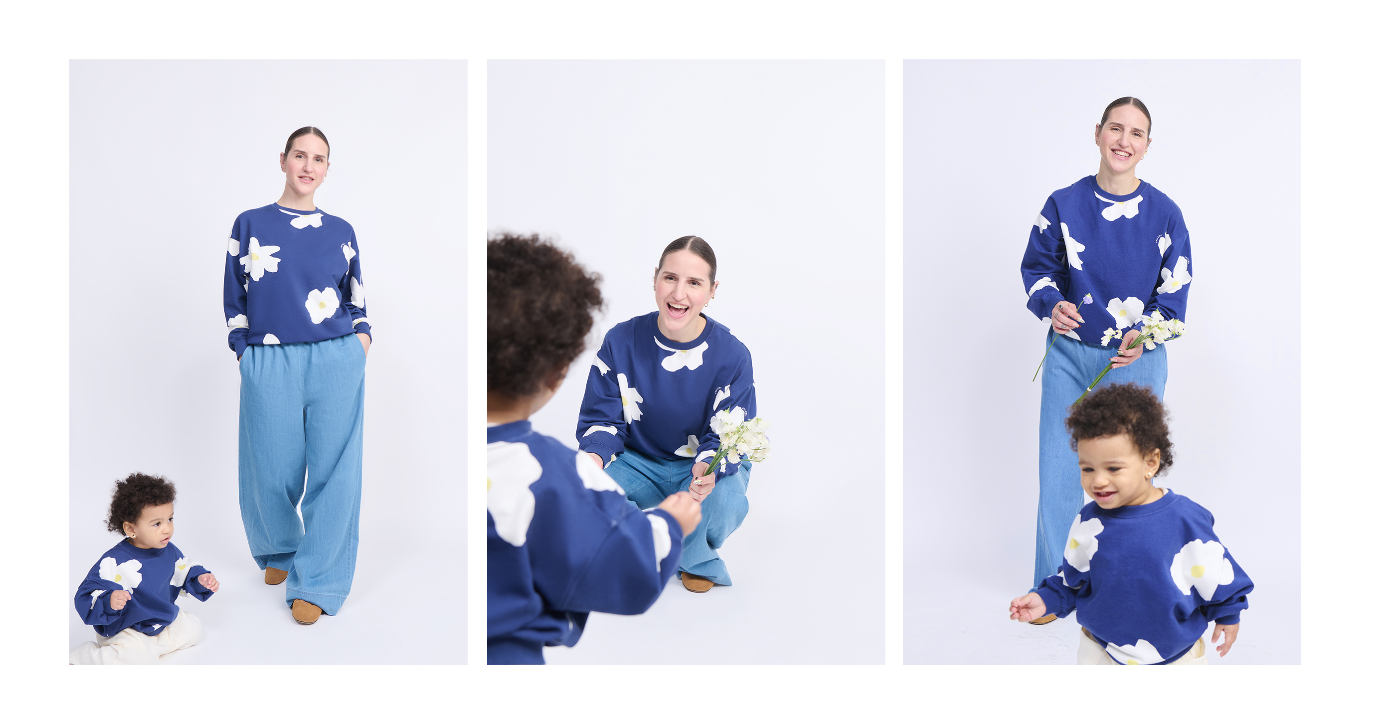 A woman and a child wearing matching blue outfits with white flowers are smiling and interacting, holding white flowers against a plain backdrop.