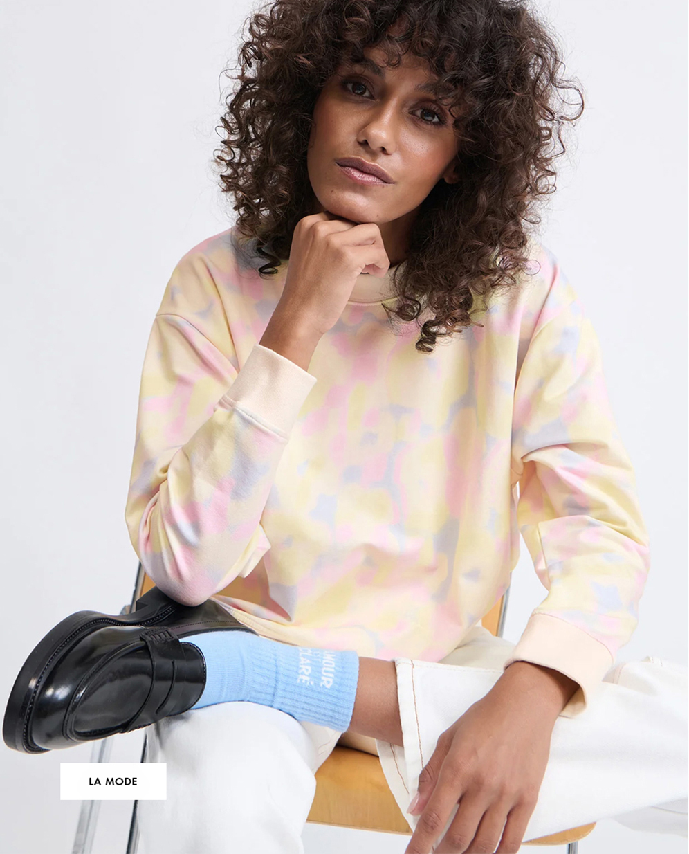 Woman in a pastel tie-dye sweatshirt and white pants sits on a chair, with one foot up, wearing blue socks and black shoes.