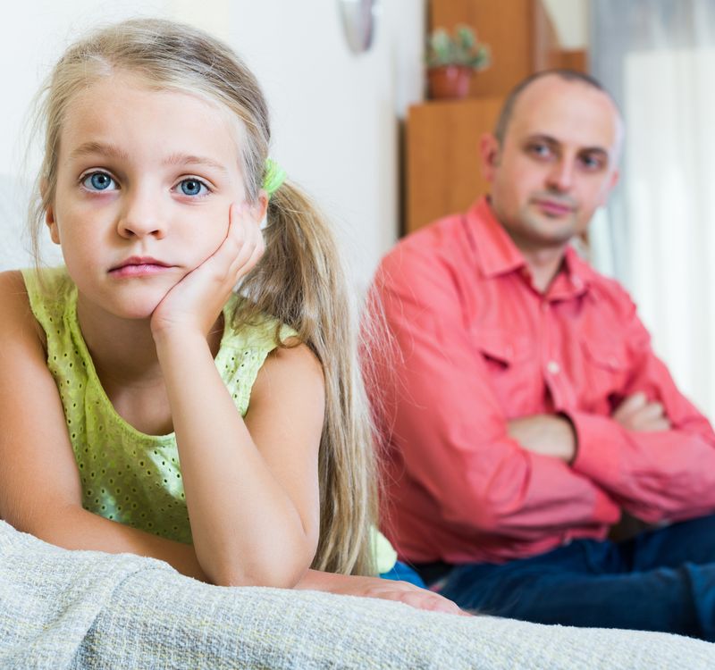 Mädchen mit kritischem Blick in die Kamera, Vater sitzt im Hintergrund und schaut sie an.