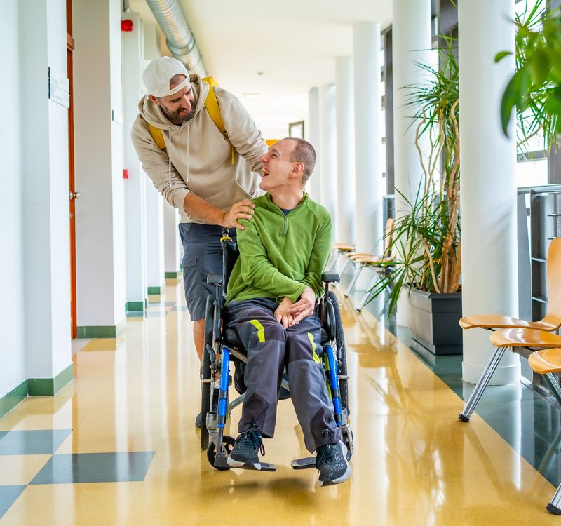 Ein Mann schiebt einen anderen Mann im Rollstuhl und sie lächeln sich an.