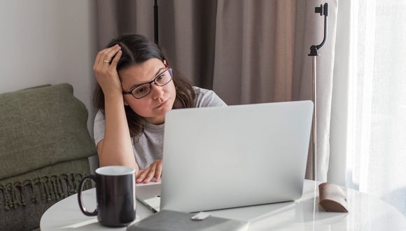 Jüngere Frau sitzt am Wohnzimmertisch und schaut mit besorgtem Blick auf den Bildschirm ihres Laptops.