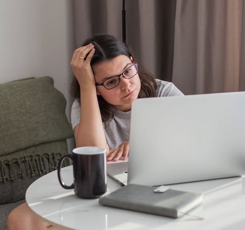 Jüngere Frau sitzt am Wohnzimmertisch und schaut mit besorgtem Blick auf den Bildschirm ihres Laptops.