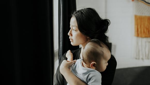 Eine junge Mutter mit Baby auf dem Arm schaut abwesend aus dem Fenster