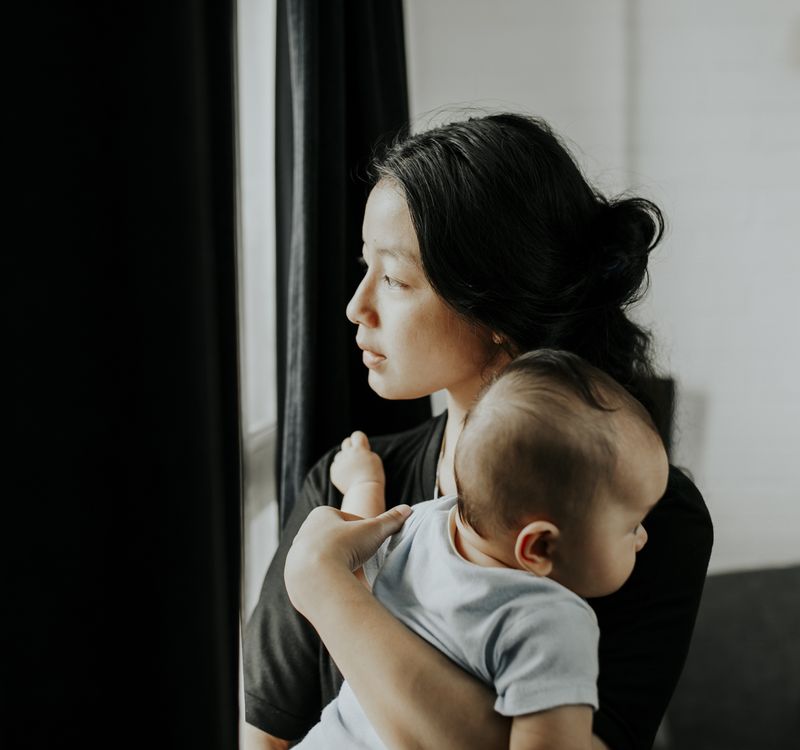 Eine junge Mutter mit Baby auf dem Arm schaut abwesend aus dem Fenster