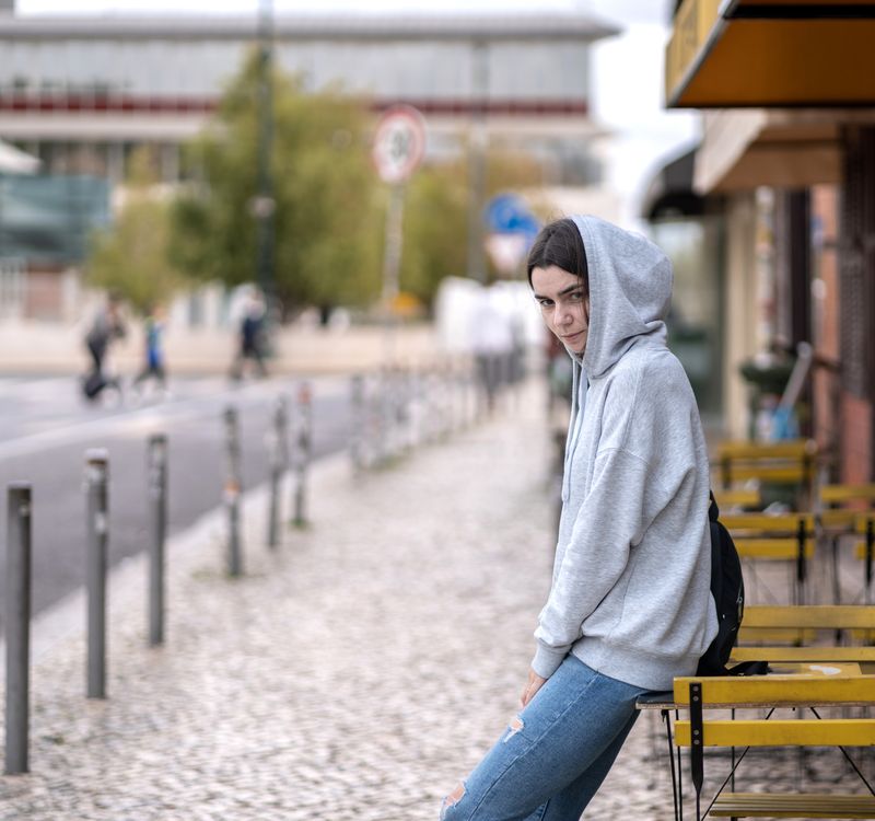 Junge Frau lehnt sich an den Tisch eines Strassencafés und schaut abwartend in die Kamera.