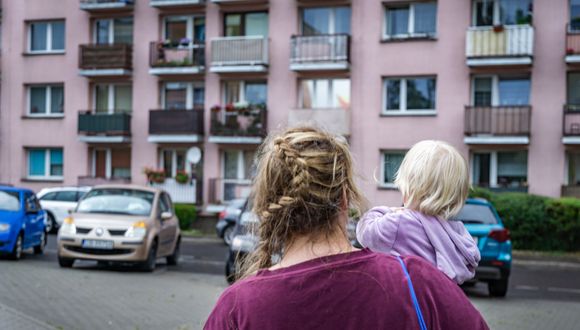 Frau mit Kind auf dem Arm steht vor einem Wohnblock.