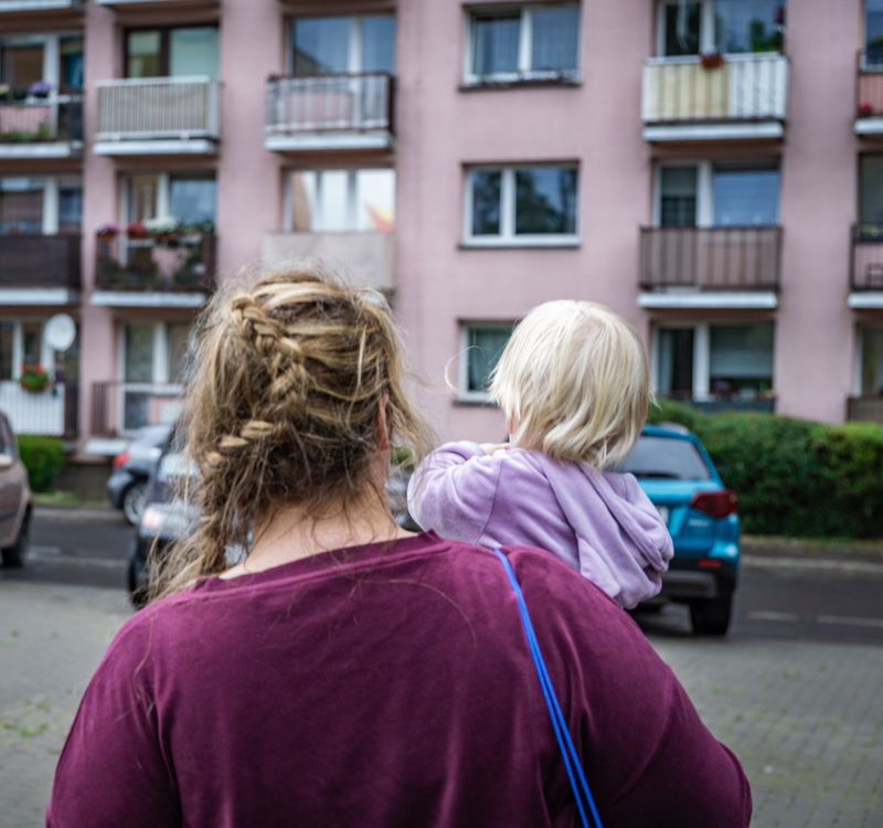 Frau mit Kind auf dem Arm steht vor einem Wohnblock.