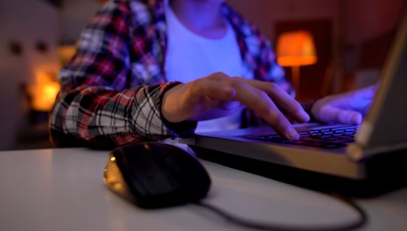 Jüngere männliche Person sitzt am Tisch und gibt etwas in die Tastatur des Laptops ein.