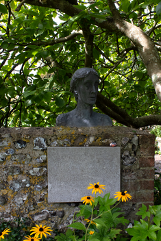 a bust of Virginia Woolf sits on top of a flint and brick wall with a commemorative plaque, surrounded by a tree and yellow flowers