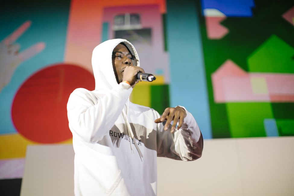 a young man of African heritage wearing a white hoodie. He is holding a microphone and performing. Behind him are some colourful shapes on the wall