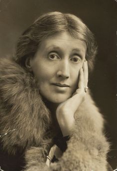 an older white woman with brown hair parted and swept back. She rests her chin in her hand and is wearing a ring and a fur coat