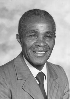a black and white photographic portrait of an older man of Caribbean heritage with short hair and a moustache. He is wearing a grey suit, white shirt and black tie.