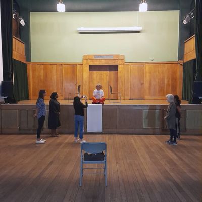 5 people stand on a wooden floor infront of an empty chair. There is a large piece of paper on the edge of the stage and a person sitting cross legged on the stage