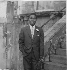 a black and white photograph of a young man of Caribbean heritage wearing a black suit, white shirt, striped tie. There is a white flower in his breast pocket. He is standing at the bottom of the stairs outside a Victorian house