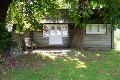 the exterior of a cabin with white wooden doors and a large tree. A bench sits on a brick patio next to the cabin surrounded by grass