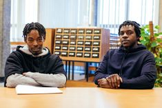 two young men of African heritage are seated at a desk. In the background is an index card set of drawers