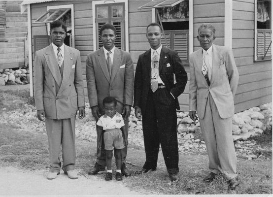 a black and white photograph of three young men, one older man and a small child of Caribbean heritage. They are wearing suits and standing in front of a wooden building