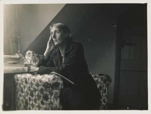 a white older woman seated, gazing outwards. She is wearing her hair behind her ears and a dark suit.