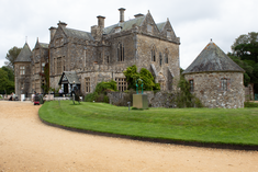 large stone house with pointed gable roof. Large stone windows. Grass bank and gravel driveway