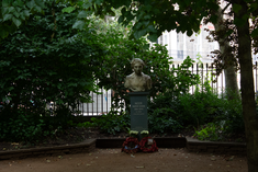 bust of Noor Inayat Khan on a stone plinth surrounded by overhanging trees and greenery. memorial poppy wreaths lie at the bottom of the plinth