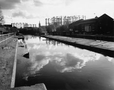 Black and white image of a canal reflecting clouds, with industrial buildings and gas holders in the background.