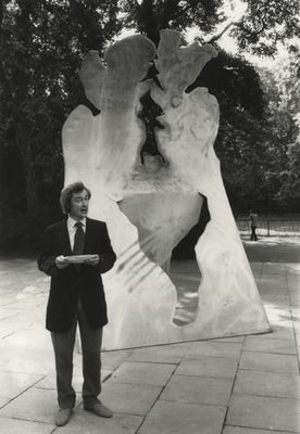 black and white image of a man wearing a suit holding a piece of paper and making a speech. Behind him is a large metal abstract sculpture.
