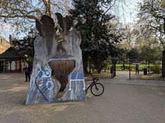 large metal sculpture covered in graffiti, wiht a bike leaning against it. The sculpture is sited on a pavement infront of park gates ad trees.