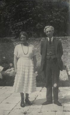 a woung white woman with dark hair parted and swept back wearing a white muslin frilled dress stands next to an older white man with grey hair and a dark suit. They are standing in front of a wall in a garden