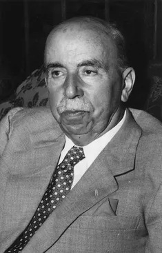 black and white image of a balding man with a moustache sitting in a chair. He is wearing a grey suit and spotted tie