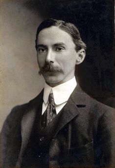 a young white man with short dark hair and a moustache is dressed in a dark suit with a stiff white collar