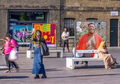 colourful image of some photographic display boards outside in Granary Square King's Cross. three people are seen in the image standing, one sitting. One of the photo panels displays an image of a man smiling wearing a red jacket