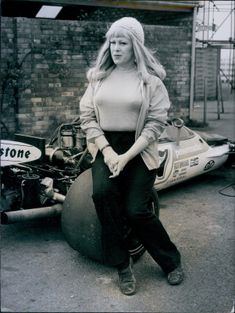 Woman in a casual outfit leans against a vintage race car tire in an industrial setting, with a brick wall backdrop and racing decals visible.