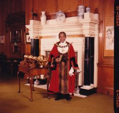 a colour photo of an older man of Caribbean heritage wearing a red, fur lined Mayor's cloak and golden Mayor's chain. He is standing in Camden's Mayor's Parlour in front of a large marble fireplace and next to a table. On the table are gold objects associated with the office of Mayor