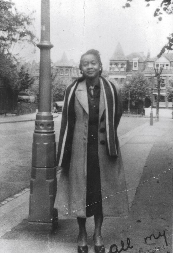 black and white image of a woman of Caribbean heritage wearing a Mac and scarf standing under a streetlight