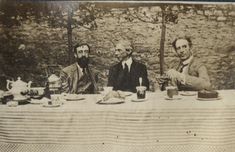 three white men seated at a long table infront of a wall in a garden. The table has a cloth, teapot, jam pot and cakes