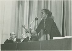 A woman passionately speaks at a podium in front of a microphone, while two men sit listening beside her on stage.