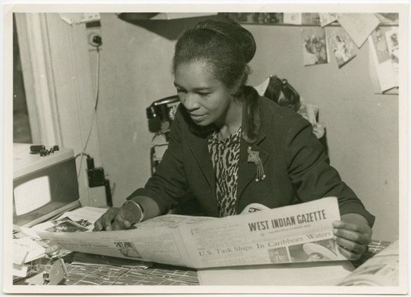 Woman reading the "West Indian Gazette" at a cluttered desk with a bulletin board behind her, appearing focused and engaged.