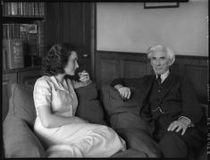 a young white woman with dark curled hair and a white dress is sitting on a sofa with a cigarette in her hand. Next to her is seated an older white man with grey hair in a dark suit. behind is a full bookcase