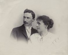 a young white man with dark hair and moustache, wearing a dark suit and white collar and cravat is seated next to a young white woman with dark hair swept into a bun. She is wearing a white dress with a large collar