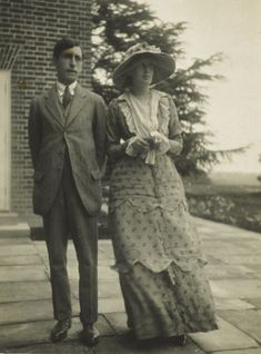 a white man with dark hair wearing a suit standing next to a white woman wearing a large hat with flowes and a long floral dress